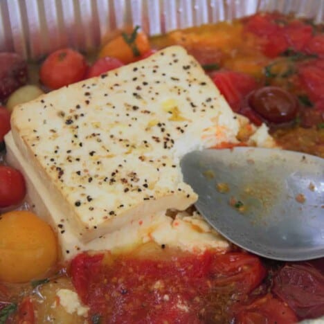 A large metal spoon gently smashes a block of cooked and seasoned feta cheese and cherry tomatoes.