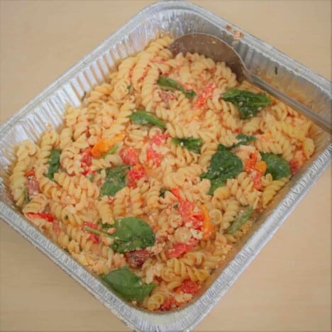 A mixture of rotini spiral pasta, cooked tomatoes, feta cheese, and fresh herbs is in a large rectangular foil pan.