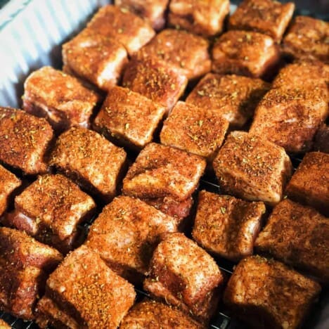 A baking rack is lined with cubes of pork belly seasoned heavily with barbecue rub.