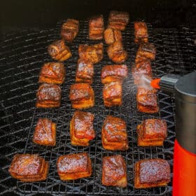 Glazed brown cubes of pork belly are spaced on a wire rack, cooking slowly in a smoker.