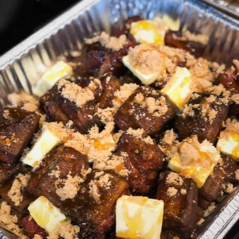 An aluminum pan holds cubes of pork belly topped with butter slices, brown sugar, and drizzled with honey.