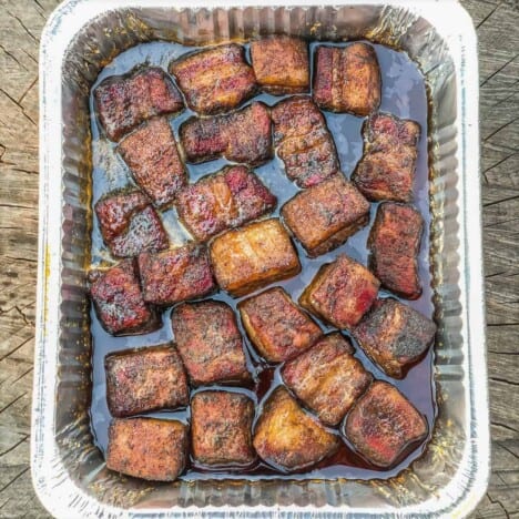 An aluminum pan is filled with smoked pork belly cubes, tossed in barbecue sauce.