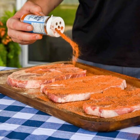 Three raw pork blade steaks are being seasoned with marinade while sitting on a cutting board with a blue and white checkered tablecloth.