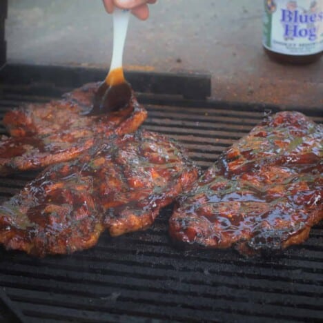 Three charred pork blade steaks are being basted with cola while sitting on a hot grill.