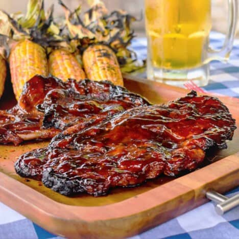 A glazed pork blade steak sits on a wooden cutting board, with grilled corn, a mug of beer, and a blue and white checkered tablecloth in the background.