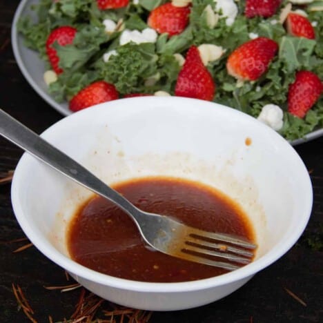Looking down into a white bowl with dressing and a fork, with a strawberry kale salad in the background.