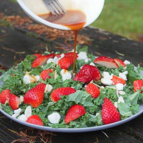 A plate of fresh kale, strawberries, and goat cheese sitting on a dark wood bench is being drizzle with salad dressing from a small white cup.