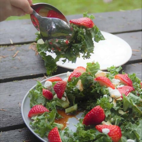 A pair of spoons is taking a serving of strawberry kale salad from a plate on a wooden picnic table, with an empty white plate in the background.