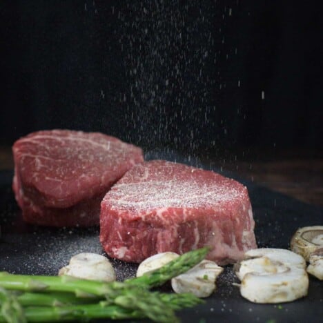 Two raw filet mignon steaks are sitting on a dark background, with fresh cooked asparagus spears in the foreground.