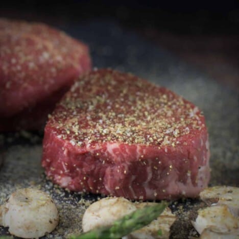 Two rare filet mignon steaks are sprinkled with herb seasoning and resting on a dark background.