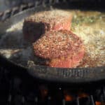 Two filet mignon steaks sit in a cast iron skillet, filled with an herb basting butter.