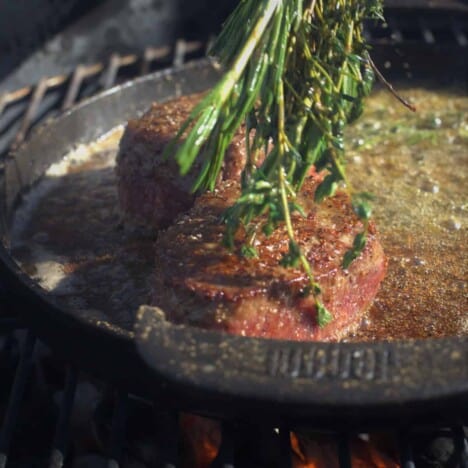 A bundle of fresh thyme is used to baste a filet mignon steak cooking in a cast iron skillet.