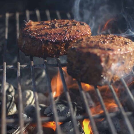 Two cooked filet mignon steaks sit on a hot grill with flames underneath.