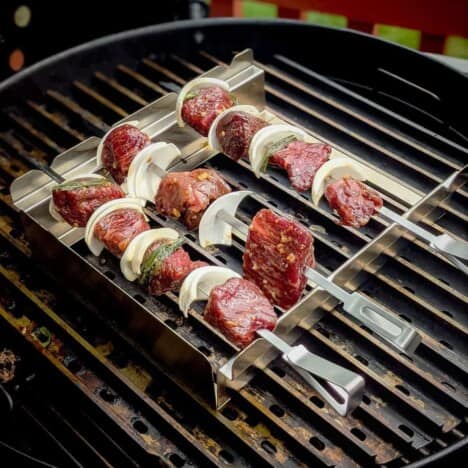 Looking down onto a hot grill with three beef, onion, and bell pepper kabobs resting on the grates.