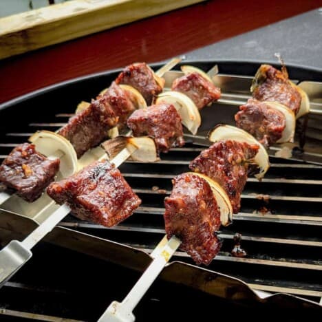 Looking across the grates of a grill where three beef, onion, and bell pepper kabobs rest.