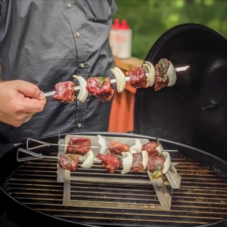 A man in a grey shirt holds a raw meat and pepper kabob over a small grill.