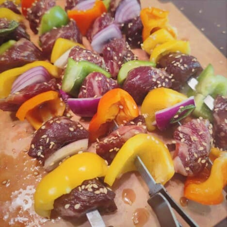 A pile of grilled beef and colorful bell pepper kabobs rests on a wooden cutting board.
