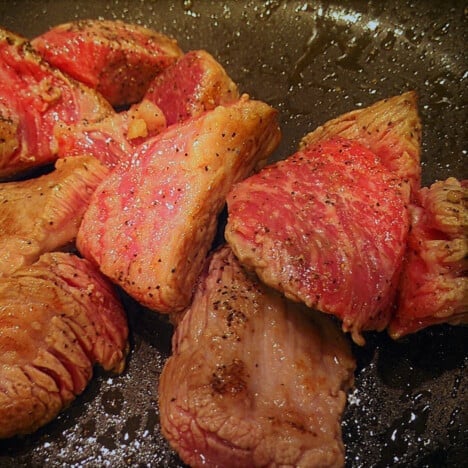 Chunks of beef being cooked in a fry pan with some edges brown but others still pink.