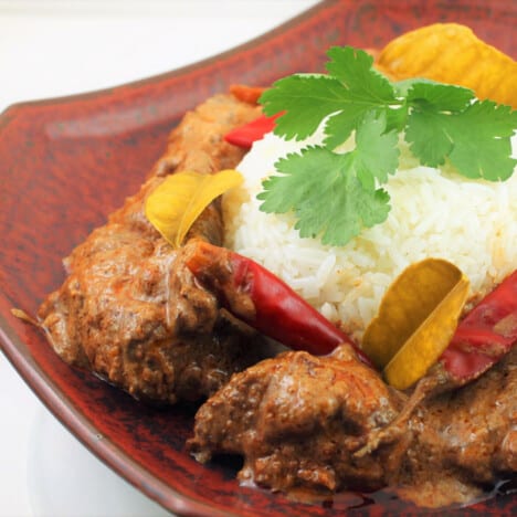 A res swuare orental plate with a mound of rice surounded by beef rendang and garnished with cilantro, red peppers a kiffir leaves.