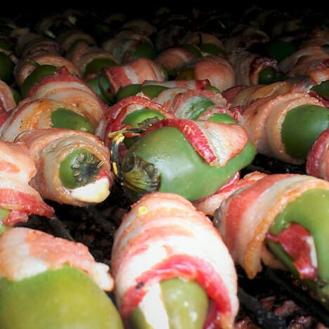 Green stuffed janapeno peppers wrapped in bacon cooking on a grill.
