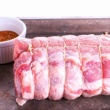 A perfectly cylindrical trussed piece of rolled pork witting on a chopping board next to a small bowl of rub.