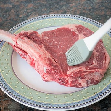 A pastry brush brushing a raw tomahawk steak on a plate.