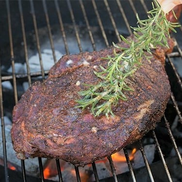 A sprig of rosemary brushing a grilled tomahawk steak on a grill grate.