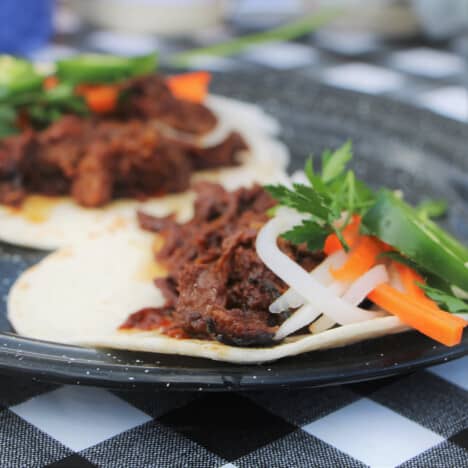 Two korean brisket tacos sitting on a black camp plate on a black and white checkered table cloth.