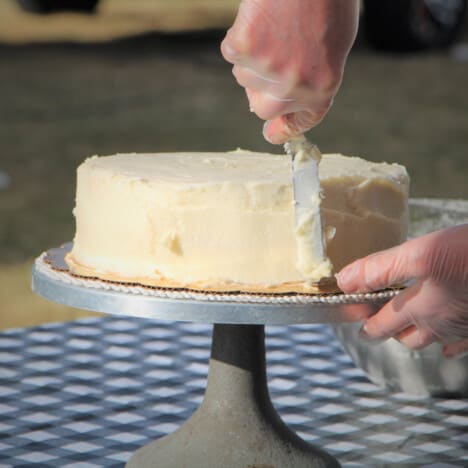 A cake on a cake stand having the frosting smoothed out with a spatula.