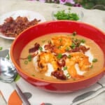 Shrimp and grits in a red bowl with crumbled bacon and sliced green onions in the background.