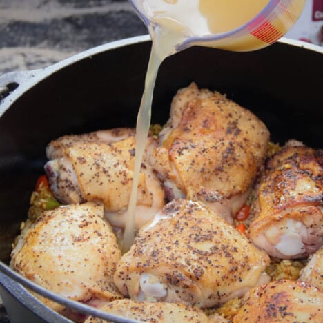 Chicken stock being poured onto fried chiken sitting on rice in a Dutch oven.