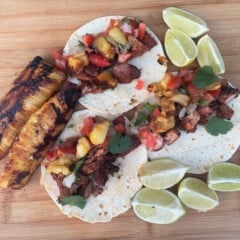 Looking down on a wooden chopping board with three tacos, grilled pineapple, and lemon wedges.