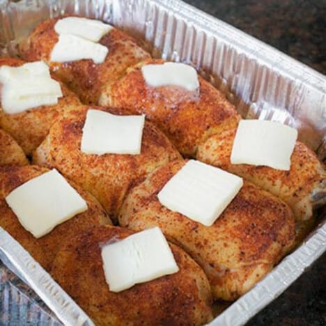 Pieces of rubbed chicken in a foil half pan, each with a pice of butter sitting on top of them.