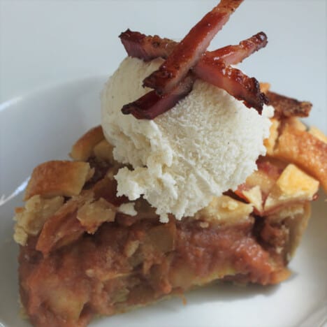 A slice of bacon apple lattice pie on a white plate topped with vanilla ice cream and bacon bits.