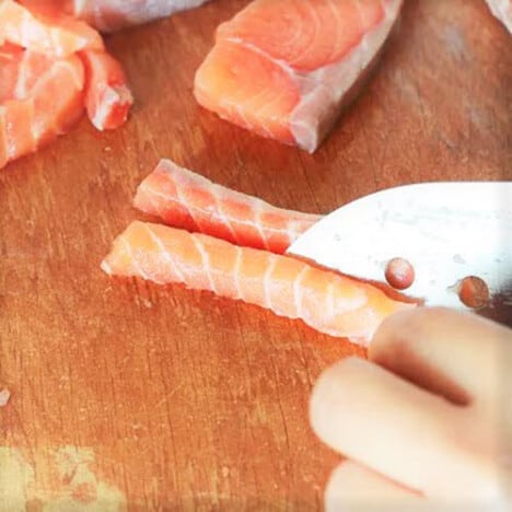 A piece of salmon is being cut with a knife on a wooden cutting board.