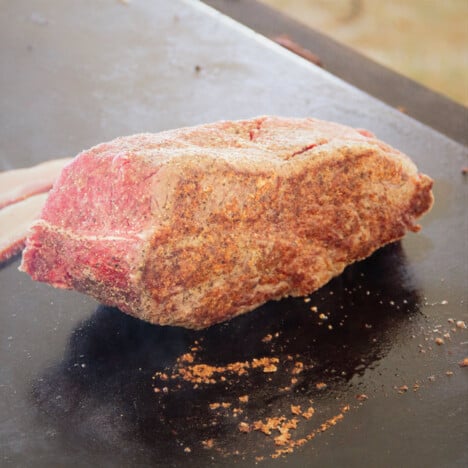 A piece of raw meat covered in rub being seared on a flat top grill.