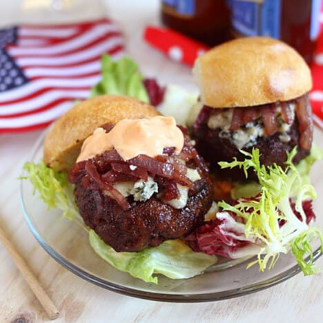 Two burgers with blue cheese, onion jam, and spicy mayonnaise sit on a glass plate with an American flag in the background.