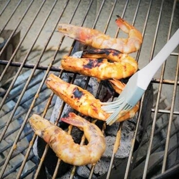 A grill brush is basting four shrimp on a grill.