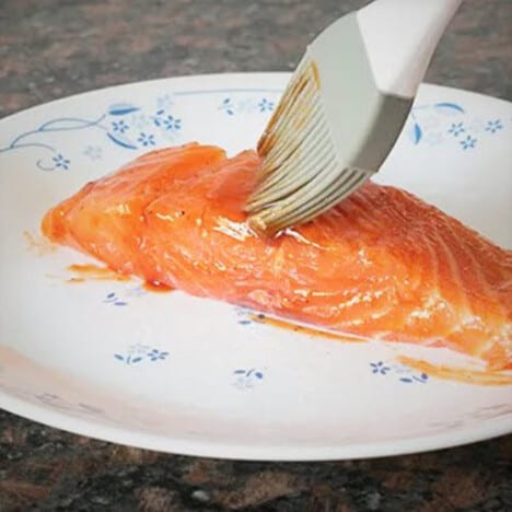 A plastic kitchen brush painting BBQ sauce onto a fillet of salmon.