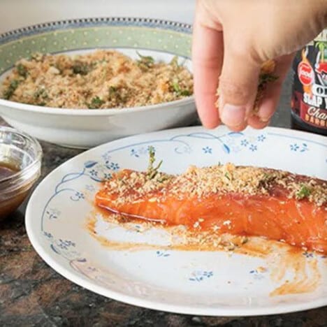 Crushed crackers being sprinkled over a fillet of salmon sitting on a white plate.