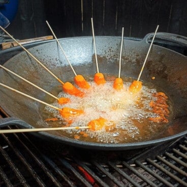 A cast iron wok over fire cooking skewered carrots.