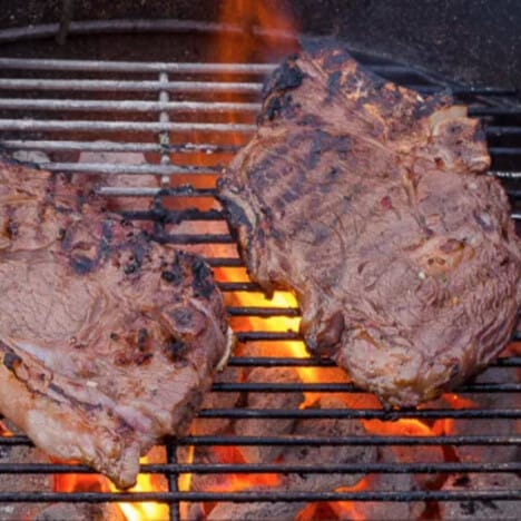 Two charred rib-eye steaks on grill grates with hot charcoals underneath.