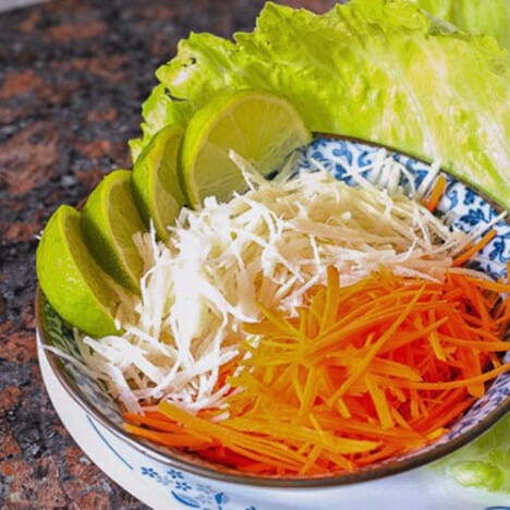 A bowl of shredded carrots and jicama are accompanied by lime wedges and lettuce leaves.