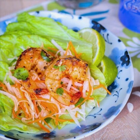 Grilled shrimp are nestled in lettuce leaves, topped with shredded carrots and jicama, with lime wedges on the side.