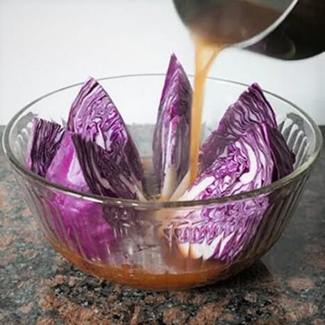 A glass bowl with wedges of purple cabbage in it and the hot brine being poured over them.