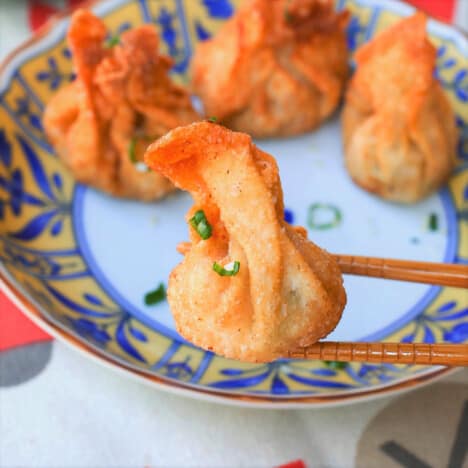 A close up of a golden fried wonton being help up by chopsticks, with a colorful plate in the background.