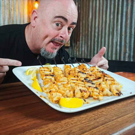 A tray of cooked calamari skewers on a white tray garnished with lemon is presented comically by Dean Schumann.