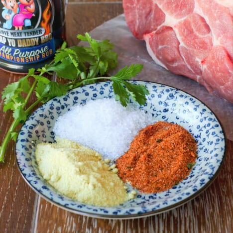 Piles of salt, garlic granules, and rub sit on blue and white plate next to a raw pork butt.