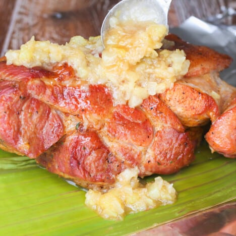 A piece of smoked pork butt lying on a banana leaf is being topped by crushed pineapple.