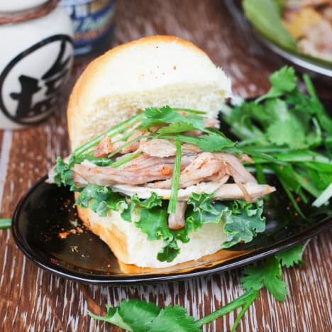 Shredded pork, fresh cilantro, and lettuce leaves are piled on a small bun resting on a black plate.